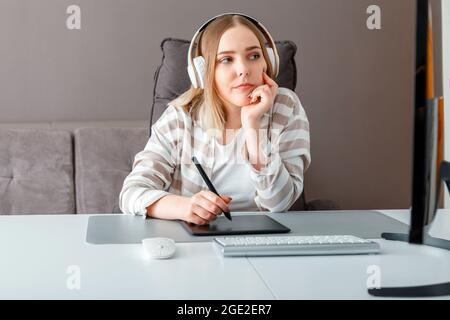 Pensive Young woman freelancer Reflects on solving problem. Designer use graphic tablet while working. Digital artist Woman in headphones works using Stock Photo