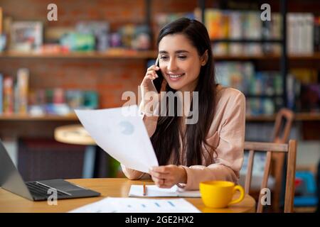 Remote office. Happy latin woman working with papers and talking with customer on cellphone, sitting at cafe interior Stock Photo