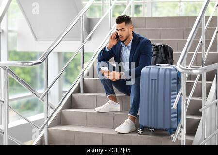 Delayed Flight. Worried Man Sitting On Stairs In Airport, Talking On Cellphone Stock Photo