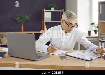 Senior male worker sitting at the table with laptop and reports taking notes Stock Photo