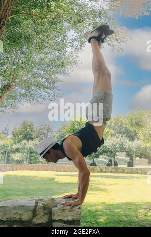 acrobatic boy with white cap performing a calisthenics handstand exercise on a stone wall with selective focus on the landscape. Stock Photo