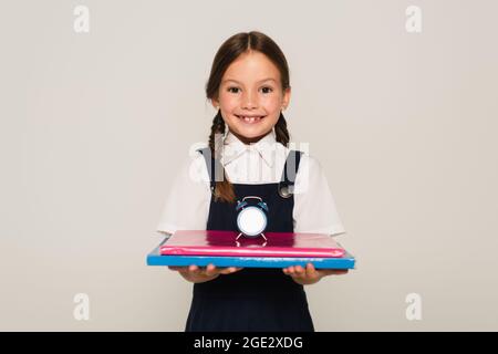 joyful schoolchild holding notebooks with small alarm clock isolated on grey Stock Photo