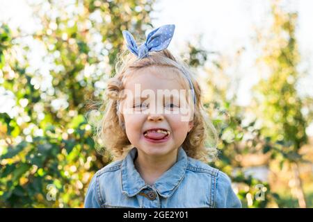 Little girl child little girl shows grimace with chewed away food and closeup portrait. Blonde caucasian child girl eating red apple and sticks out Stock Photo