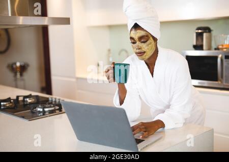 Smiling african american woman with beauty face mask, in kitchen using laptop and drinking coffee Stock Photo