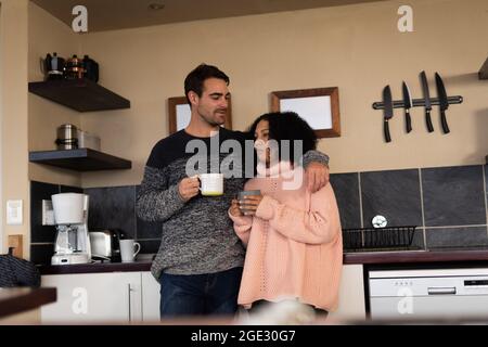 Happy diverse couple in kitchen embracing drinking coffee and smiling Stock Photo