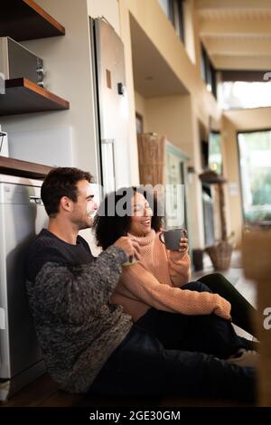 Happy diverse couple in kitchen sitting on floor and drinking coffee Stock Photo