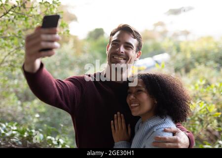 Happy diverse couple on balcony embracing taking selfie and smiling Stock Photo