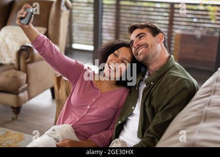 Happy diverse couple sitting on sofa in living room taking selfie and smiling Stock Photo