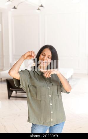 Happy excited indian woman holding keys in hand and points on it, overjoyed with buying own estate. Cheerful eastern woman relocated in new apartment, rent house. Achievement goal. Vertical shot Stock Photo