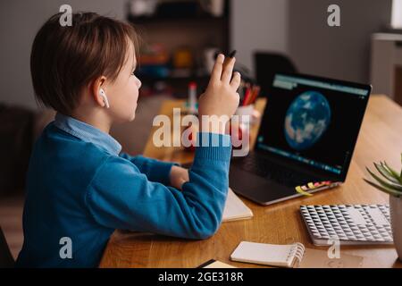 A Preteen boy uses a laptop to make online classes, globe on the screen, making notes Stock Photo