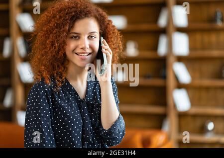 Close-up portrait of young charming woman talking on phone with partners standing and leaned on the office desk, curly red-haired businesswoman chatting on the smartphone in a relaxed atmosphere Stock Photo