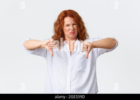 Disgusted middle-aged redhead woman, female entrepreneur grimacing dissatisfied, pointing fingers down and shaking head in rejection, dislike and Stock Photo