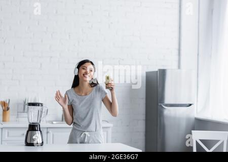 happy asian woman in headphones holding glass of smoothie and waving hand at camera near electric shaker Stock Photo