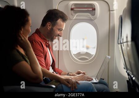 Side view of cheerful caucasian man smiling while using laptop computer, sitting on the airplane Stock Photo