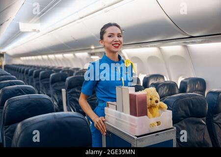 Cheerful female air hostess in bright blue uniform smiling at camera while leading trolley cart with gifts through empty plane aisle Stock Photo