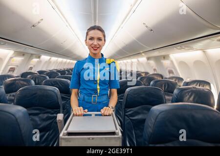 Attractive female cabin attendant in bright blue uniform smiling at camera while leading trolley cart through empty plane aisle Stock Photo