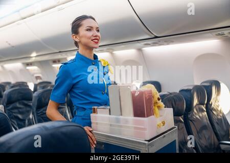 Pretty stewardess in bright blue uniform smiling aside while leading trolley cart with gifts through empty plane aisle Stock Photo