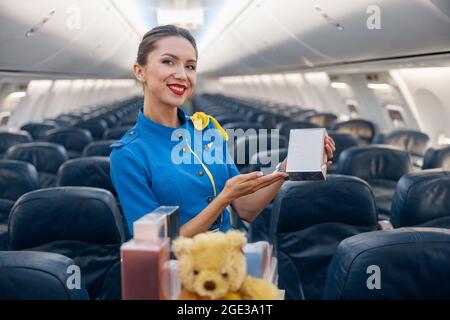 Pretty stewardess in bright blue uniform smiling at camera, advertising perfume as a gift while leading trolley cart through empty plane aisle Stock Photo