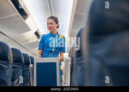 Smiling female cabin attendant leading trolley cart through empty plane aisle Stock Photo