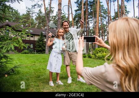Smiling father and kid holding badminton rackets near mother taking photo on smartphone outdoors Stock Photo