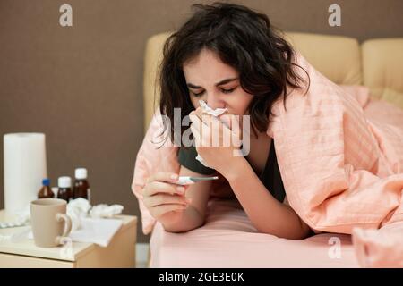 young sick woman in bed at home. woman under a blanket measures temperature with thermometer. girl having influenza symptoms Stock Photo