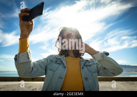 Mixed race woman taking selfie with smartphone on sunny day by seaside Stock Photo