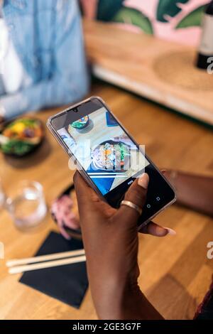 Unrecognized african woman taking a photo of her poke bowl in a restaurant. Stock Photo