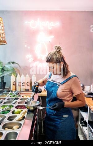 Female chef wearing apron preparing poke bowl and using kitchen blowtorch. Stock Photo
