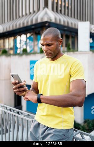 African American man with yellow t-shirt synchronizing mobile phone and smartwatch to do sports in the city. Stock Photo