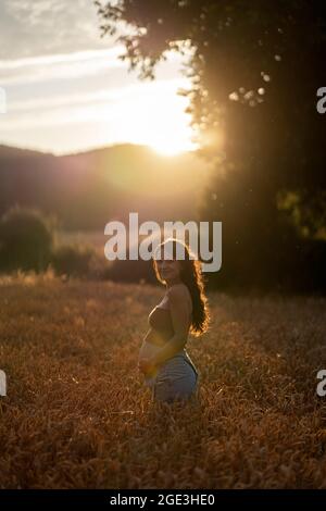 Pregnant woman smiling and standing in wheat field. She's touching her belly and looking at camera.