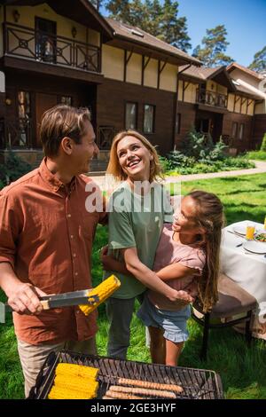 Man embracing smiling family while preparing corn on grill outdoors Stock Photo