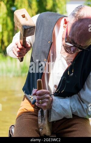 Close up of a senior man, Medieval carpenter living history reenactor, using mallet and chisel to shape some wood with green foliage background. Stock Photo