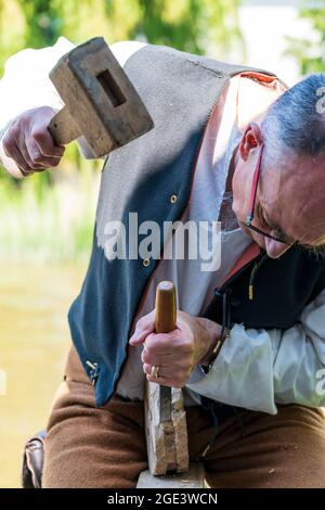 Close up of a senior man, Medieval carpenter living history reenactor, using mallet and chisel to shape some wood with green foliage background. Stock Photo
