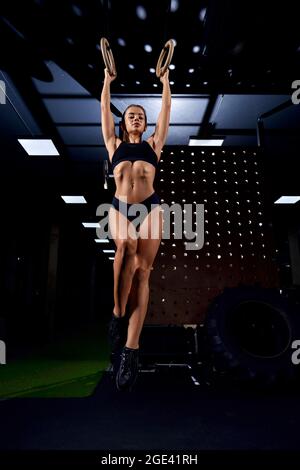 Front view of young attractive sportswoman doing ring exercise at gym. Fit female posing, doing fitness workout complex using gymnastic rings. Concept of sport, gymnastic. Stock Photo