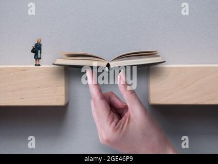 Hand supporting open book bridging the gap between wooden blocks for female miniature figure to cross Stock Photo