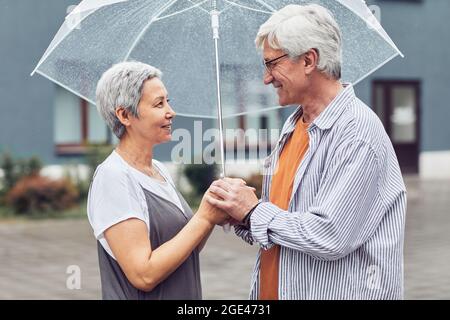 Side view portrait of modern senior couple looking at each other while standing under umbrella in city Stock Photo