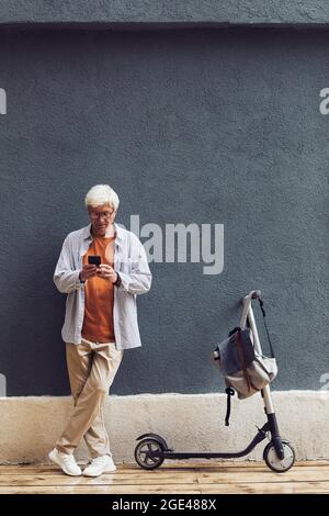 Graphic full length portrait of trendy senior man using smartphone outdoors while standing by electric scooter Stock Photo