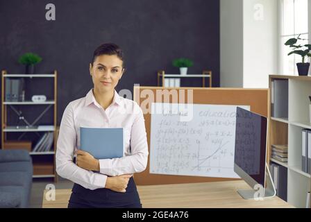 Young teacher standing in the classroom, holding a book and looking at the camera Stock Photo