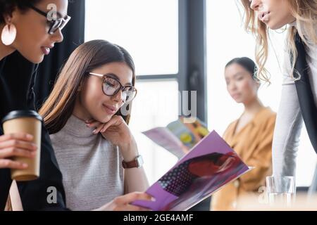 brunette businesswoman smiling while reading magazine near multiethnic coworkers Stock Photo