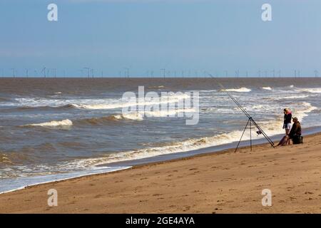 People sea fishing on the sandy beach at Chapel St Leonards in Lincolnshire England UK with offshore wind farm turbines visible on the horizon. Stock Photo