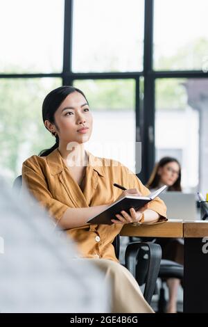 pensive asian businesswoman looking away while writing in notebook on blurred foreground Stock Photo