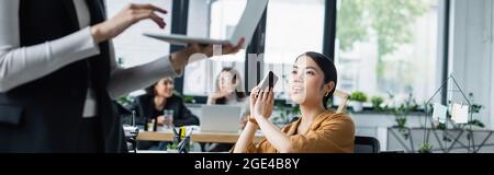 asian businesswoman covering smartphone with hand near colleague using laptop on blurred foreground, banner Stock Photo