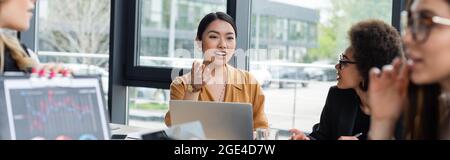 smiling asian woman pointing with hand while talking to blurred multiethnic colleagues, banner Stock Photo