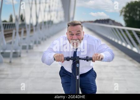 Gleeful laughing man speeding along on an electric scooter in a close up view as he approaches the camera head on while crossing a bridge Stock Photo