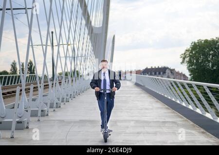 Businessman commuting in the city using an electric scooter smiling as he approaches the camera while crossing a bridge over a river at sunset Stock Photo