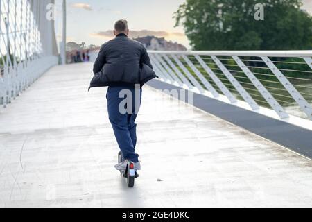 Rear view of a man riding an electric scooter on a bridge at sunset towards distant people crossing the walkway in an urban transport concept Stock Photo