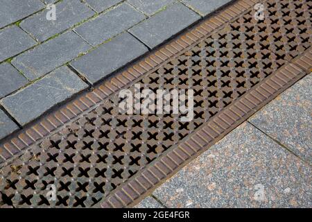 a drainage ditch covered with an iron patterned lattice on a sidewalk made of granite tiles stones close-up of a stormwater drainage system from a pav Stock Photo