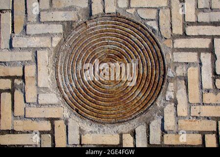 iron rusty round manhole on the sidewalk paved with stone tiles top view, old sewer hatch on a brick road. Stock Photo