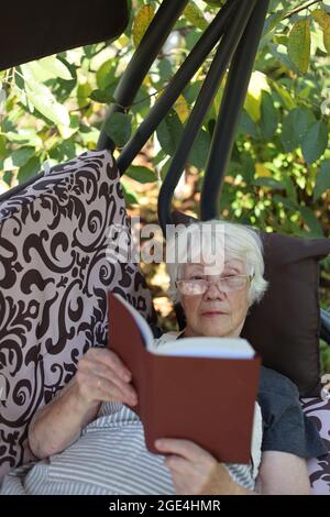 An elderly gray-haired woman lies on a garden swing and reads a book. National Book Lovers Day concept. Stock Photo