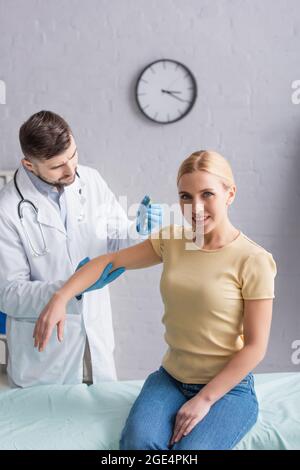 doctor in white coat and latex gloves vaccinating woman smiling at camera in clinic Stock Photo